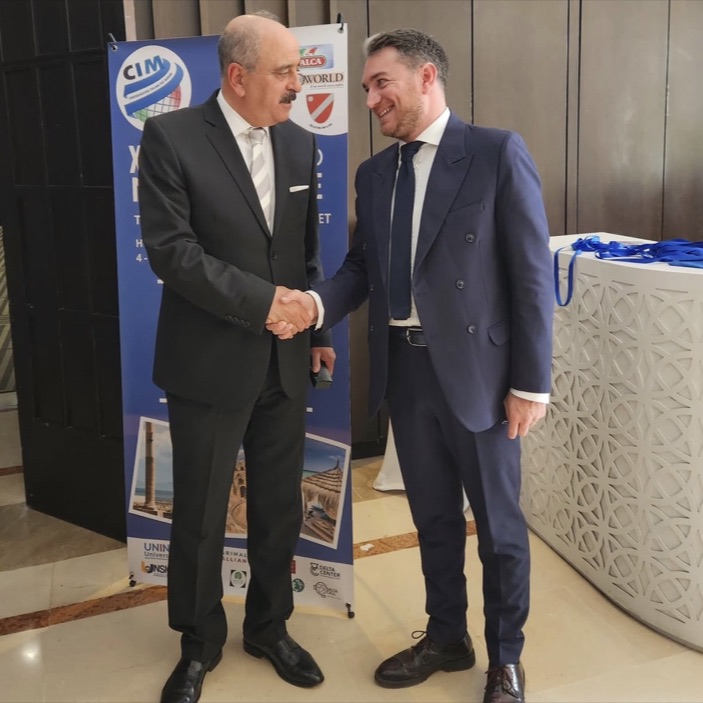 Enrico Celiberti stringe la mano a un ospite davanti al banner di un convegno